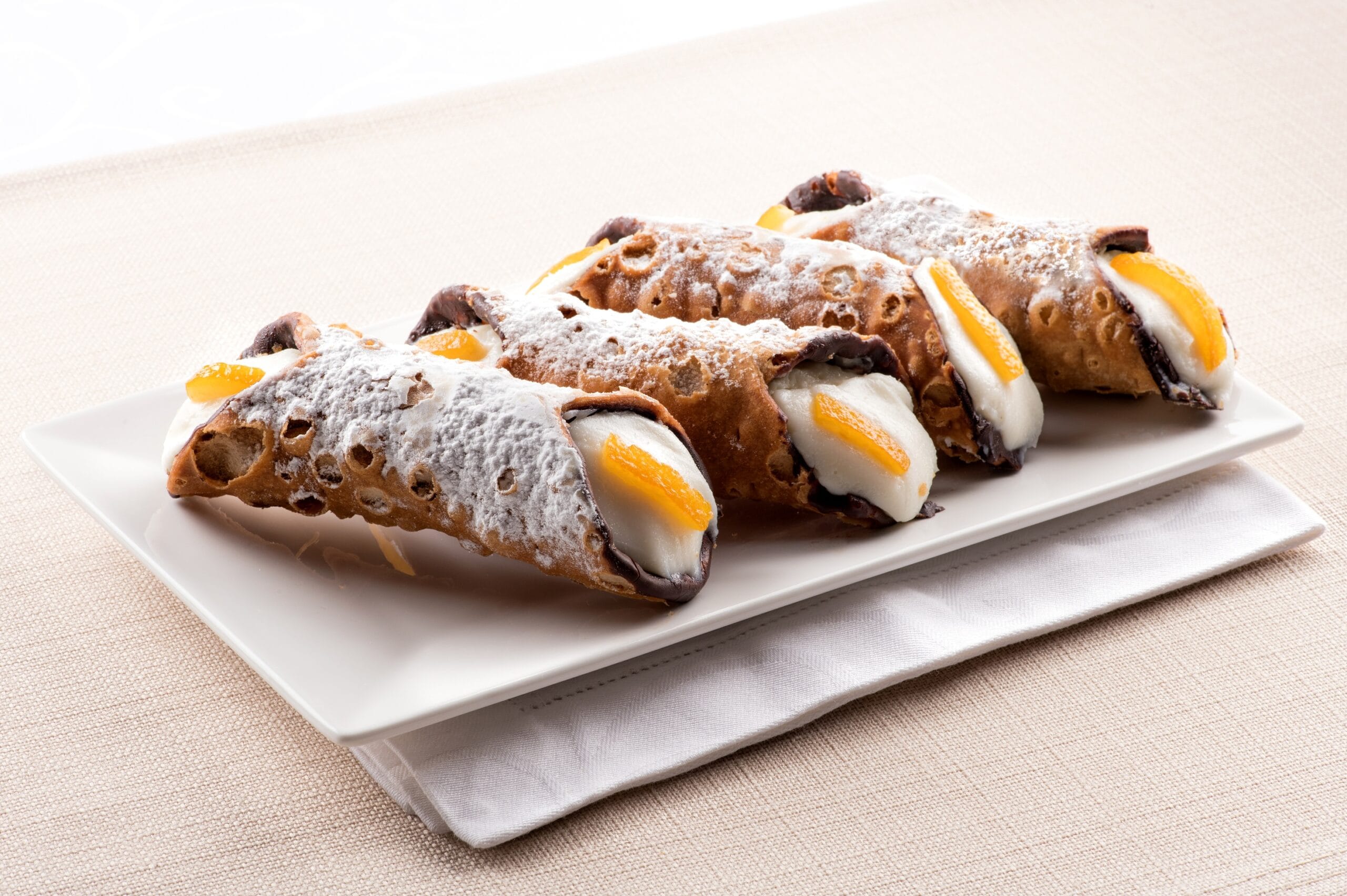 Four cannoli Sicilia, or Sicilian cannoli, deep fried pastry tubes with a sweet ricotta filling garnished with colorful orange rind and served on a rectangular plate